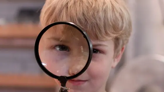 Ein Kind mit blonden Haaren hält eine Lupe vor sein Gesicht, sodass ein Auge vergrößert erscheint.