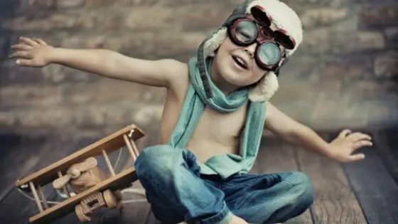 Ein kleiner Junge mit Fliegerkappe und Schutzbrille spielt auf einem Holzboden mit einem Holzflugzeug.
