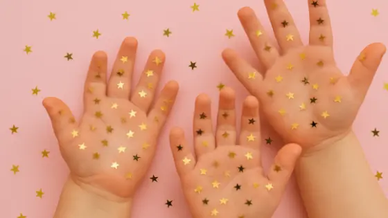 Zwei Kinderhände vor einem rosa Hintergrund, bedeckt mit goldenen Sternenkonfetti.