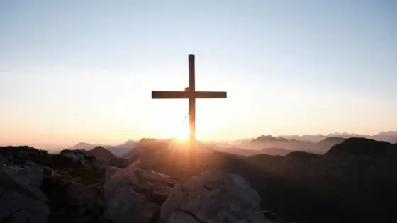 Ein Holzkreuz steht vor einer Bergkulisse während eines strahlenden Sonnenaufgangs.