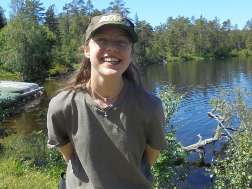 Eine junge Frau mit Brille und Cap lächelt vor einem See in einer Waldlandschaft.